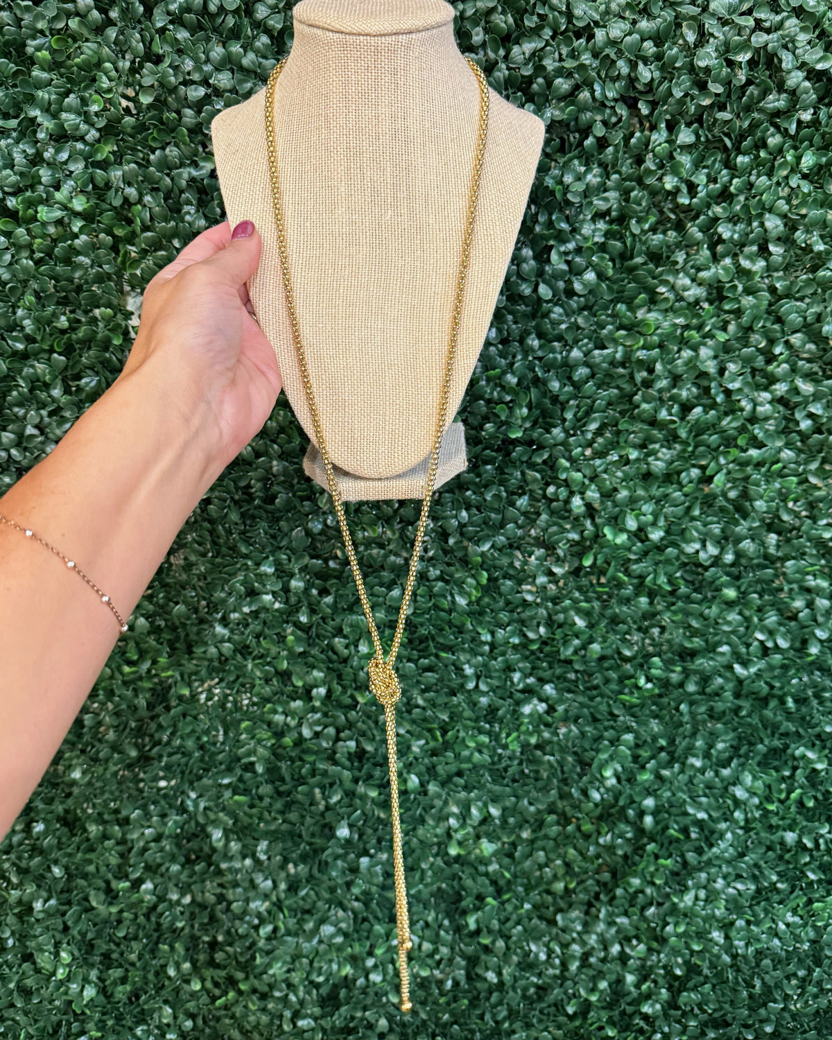 Chansuttpearls gold lariat-style necklace with a knotted center detail is displayed on a beige fabric mannequin bust. A person's hand with dark nail polish is visible on the left side holding the display bust, set against a green artificial foliage background.