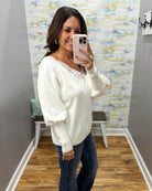 A woman takes a mirror selfie wearing a cream-colored knit sweater with lace trim at the neckline and blue jeans in a fitting room.