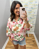 Woman taking a mirror selfie wearing a floral blouse and denim shorts.