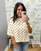 A woman taking a mirror selfie wearing a cream-colored knit sweater with brown polka dots and blue jeans, holding a light pink smartphone.