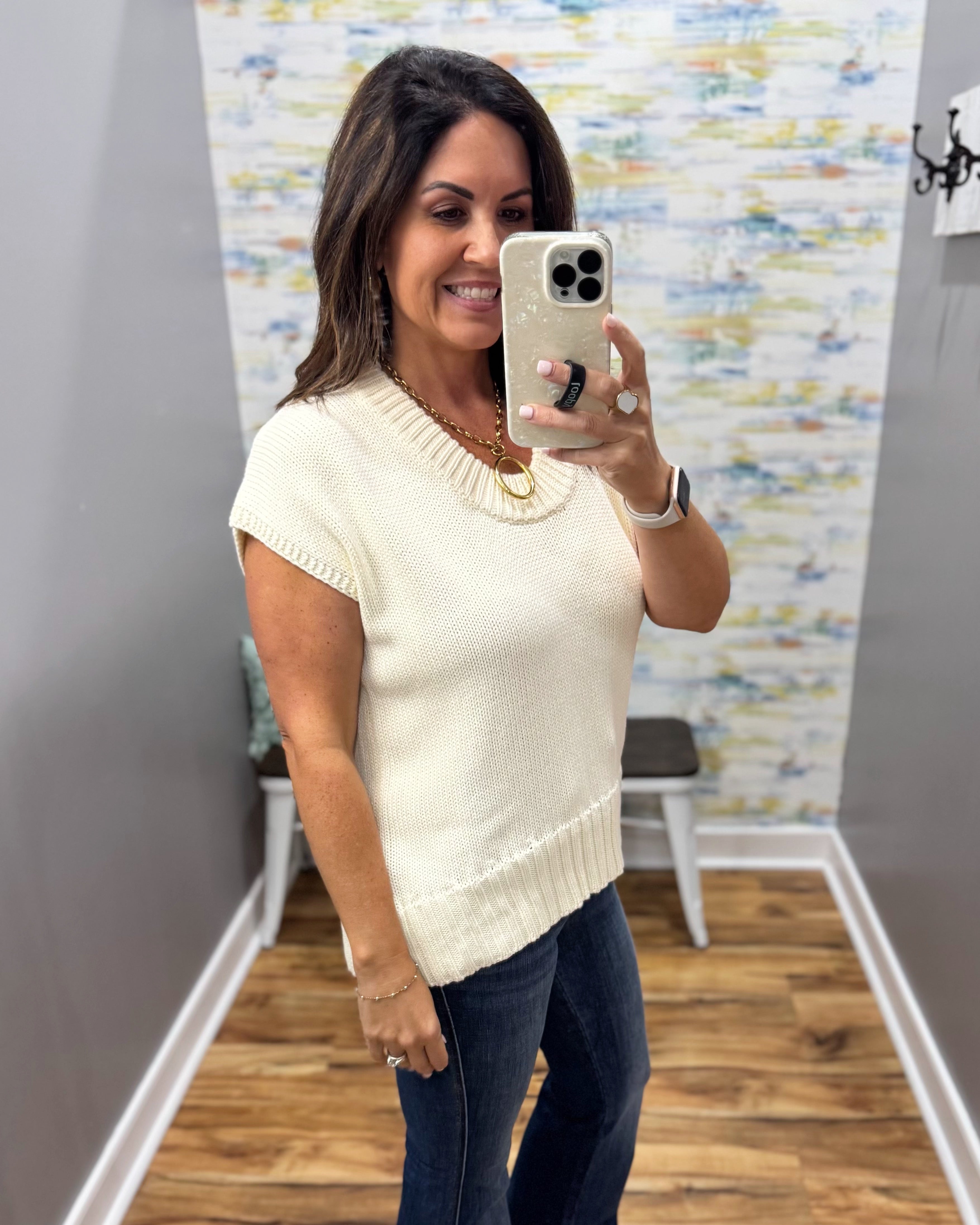 A woman takes a mirror selfie wearing a cream-colored short-sleeve knit top and dark blue jeans in a home or boutique setting with a wood floor and abstract wall art.