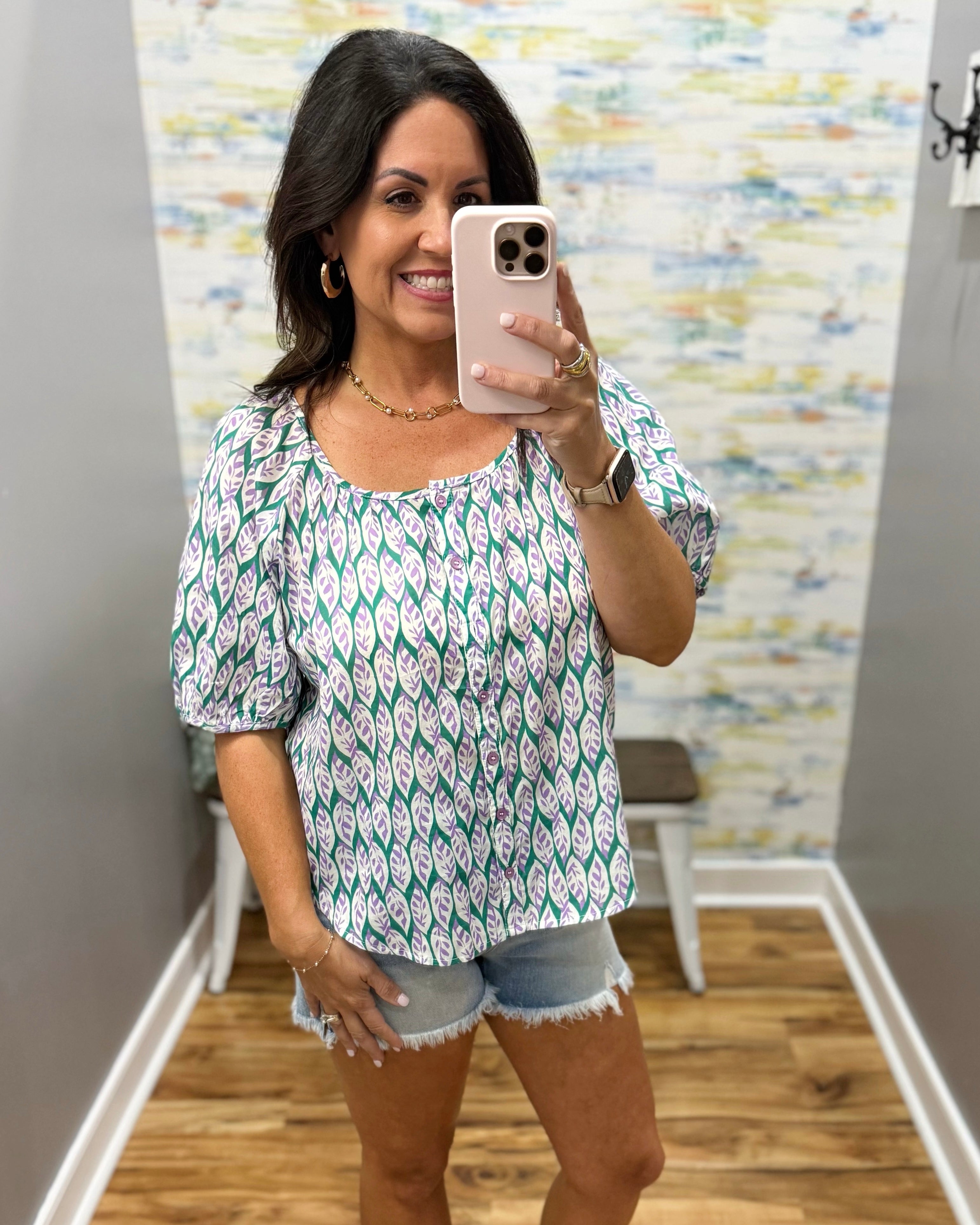 Woman taking a mirror selfie wearing a patterned top and denim shorts in a room with a textured wall.