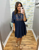 Woman taking a mirror selfie wearing a navy dress with colorful embroidery in a room with patterned wallpaper.