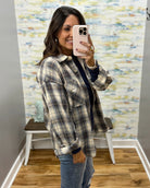 A woman takes a mirror selfie wearing an oversized plaid button-up shirt in beige, brown, and navy, layered over a dark top and paired with blue jeans.