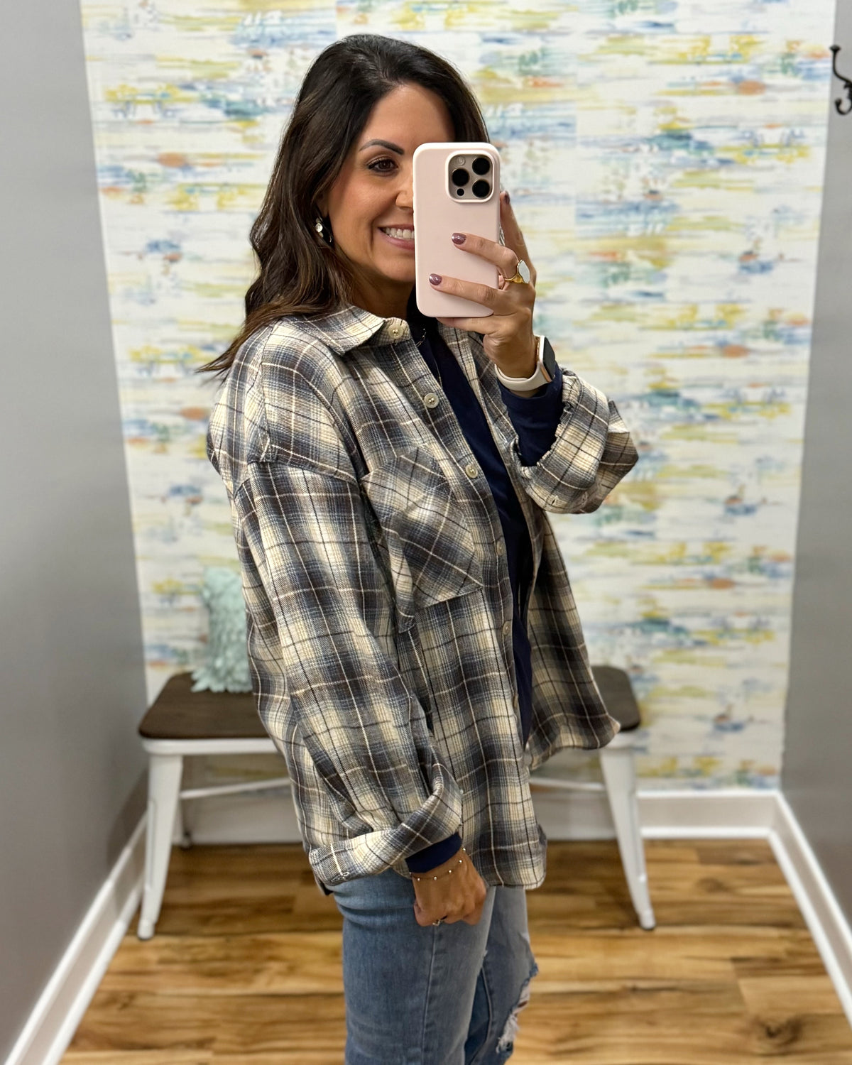 A woman takes a mirror selfie wearing an oversized plaid button-up shirt in beige, brown, and navy, layered over a dark top and paired with blue jeans.