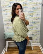 A woman takes a mirror selfie wearing an olive green ruffled blouse and dark blue jeans in front of a patterned wallpaper background.