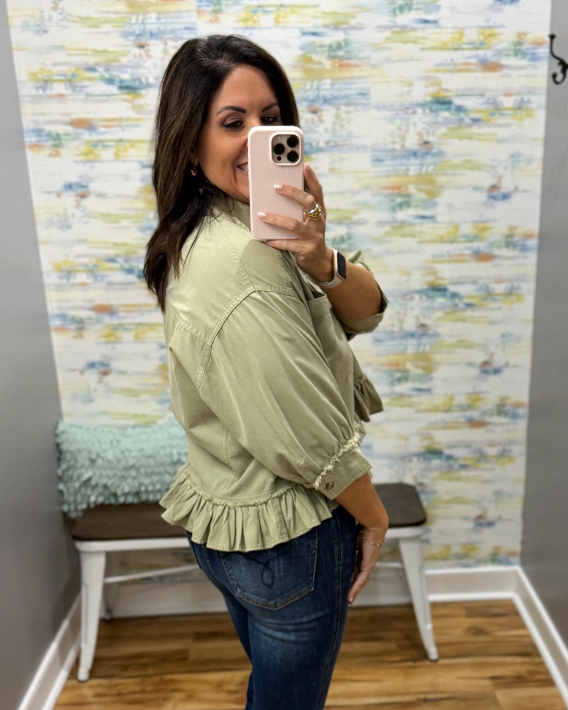 A woman takes a mirror selfie wearing an olive green ruffled blouse and dark blue jeans in front of a patterned wallpaper background.