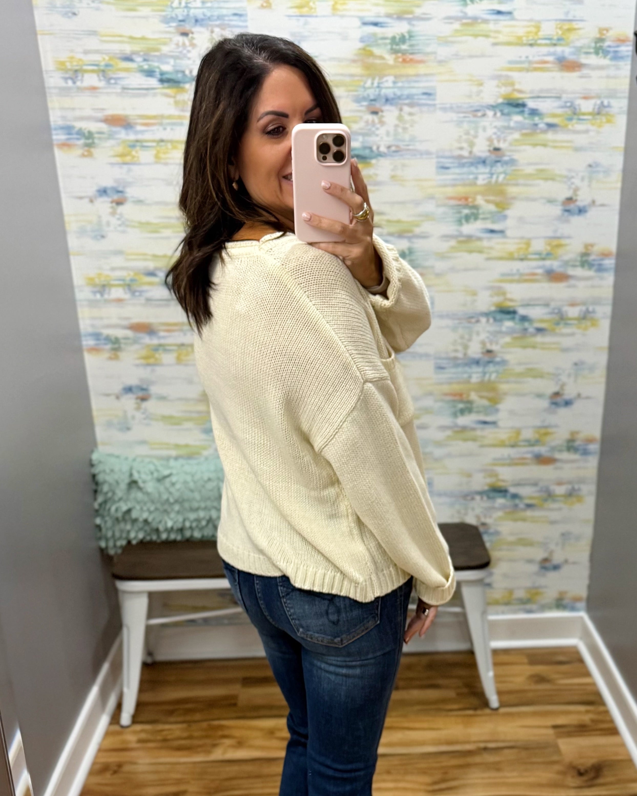 A woman takes a mirror selfie in a fitting room wearing a cream-colored knit sweater and dark blue jeans.