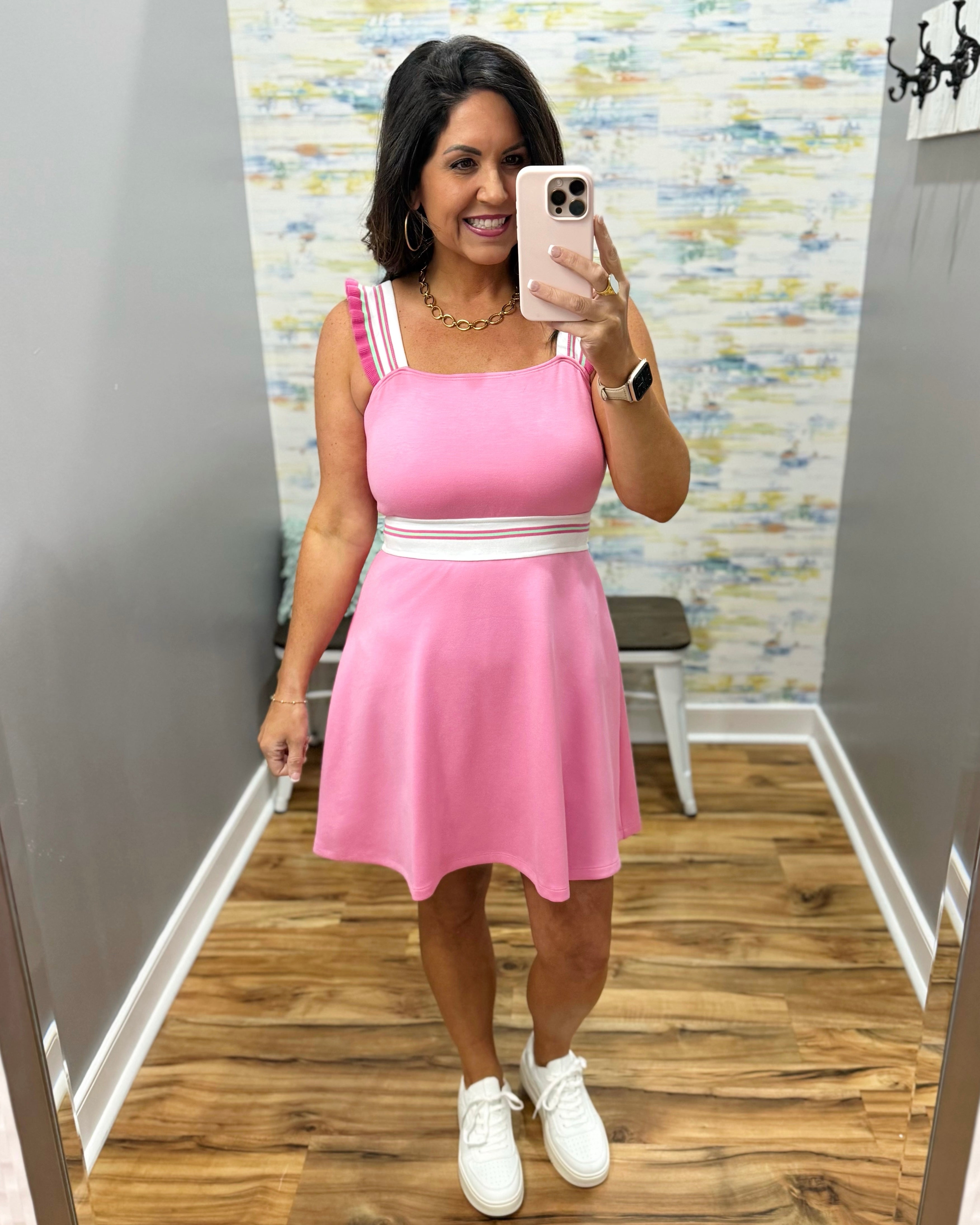 Woman taking a mirror selfie wearing a pink dress with white sneakers in a room with patterned wallpaper.