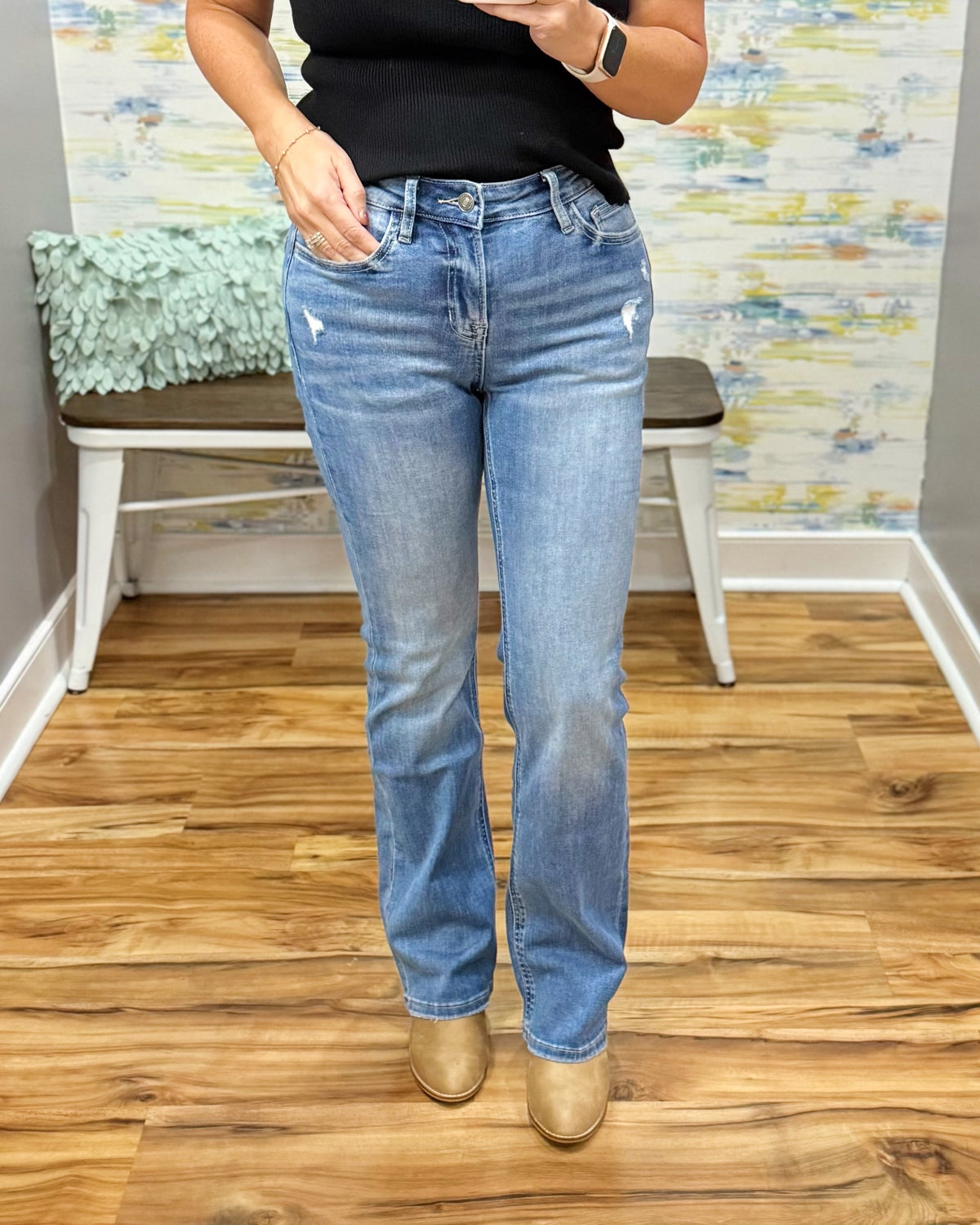 A person wearing mica bootcut jeans with subtle front distressing, paired with a black top and tan ankle boots, standing in an indoor room with wood flooring and a bench in the background.