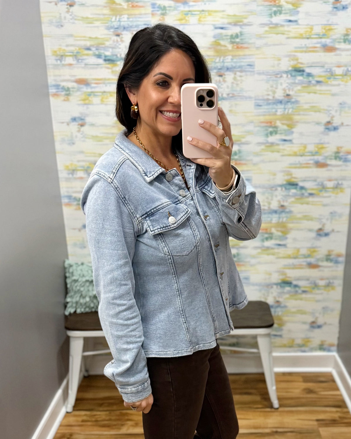 A woman takes a mirror selfie wearing a light-wash blue denim jacket and dark brown pants in a fitting room with a patterned wallpaper background.