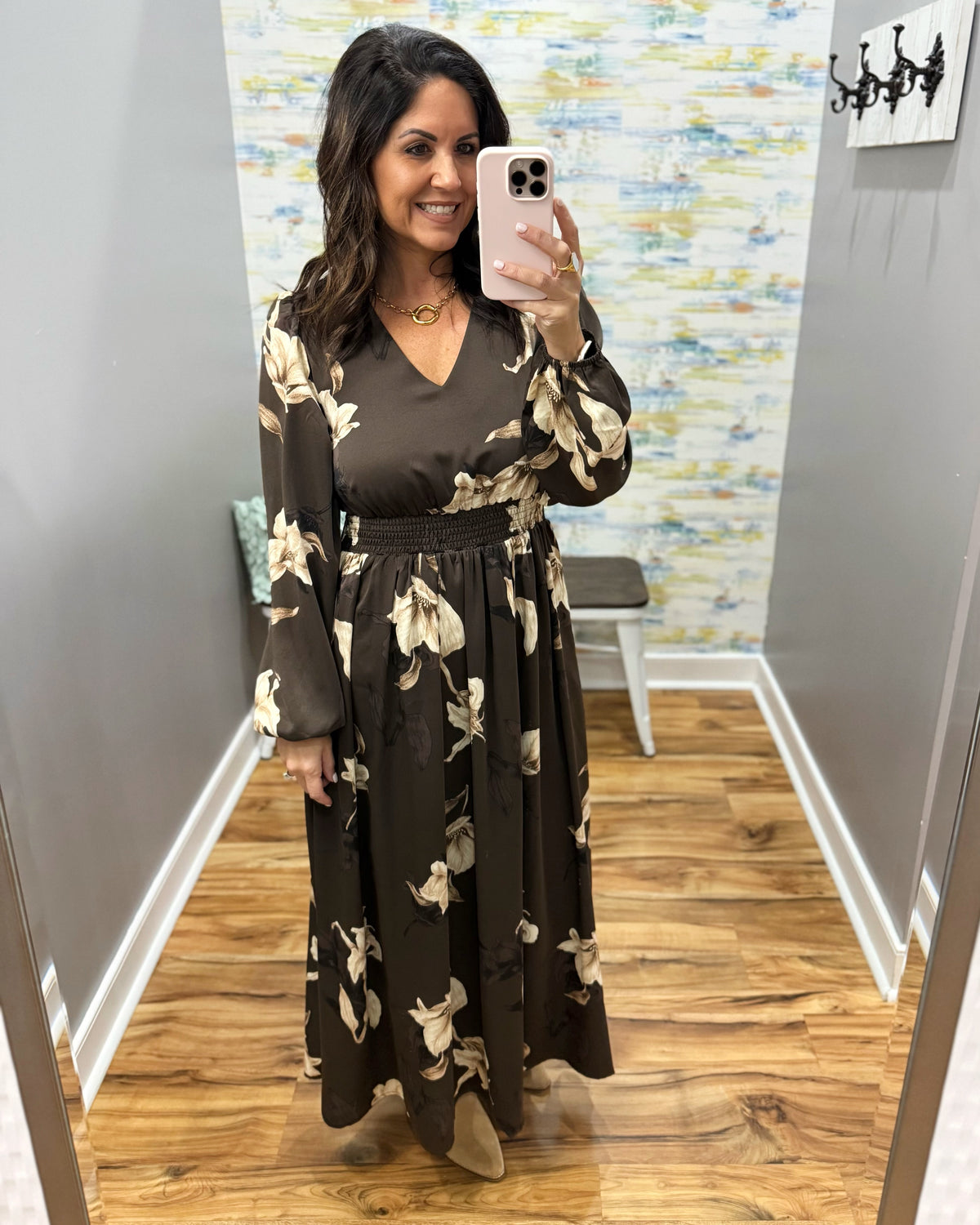 A woman takes a mirror selfie in a fitting room wearing a dark brown long-sleeve maxi dress with a beige floral pattern and a matching waist tie belt.