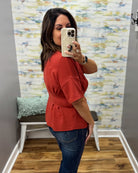 A woman takes a mirror selfie in a fitting room wearing a rust-orange short-sleeve peplum top and dark blue jeans.