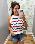 Woman taking a mirror selfie wearing a white sleeveless top with red and blue wavy patterns and denim shorts.