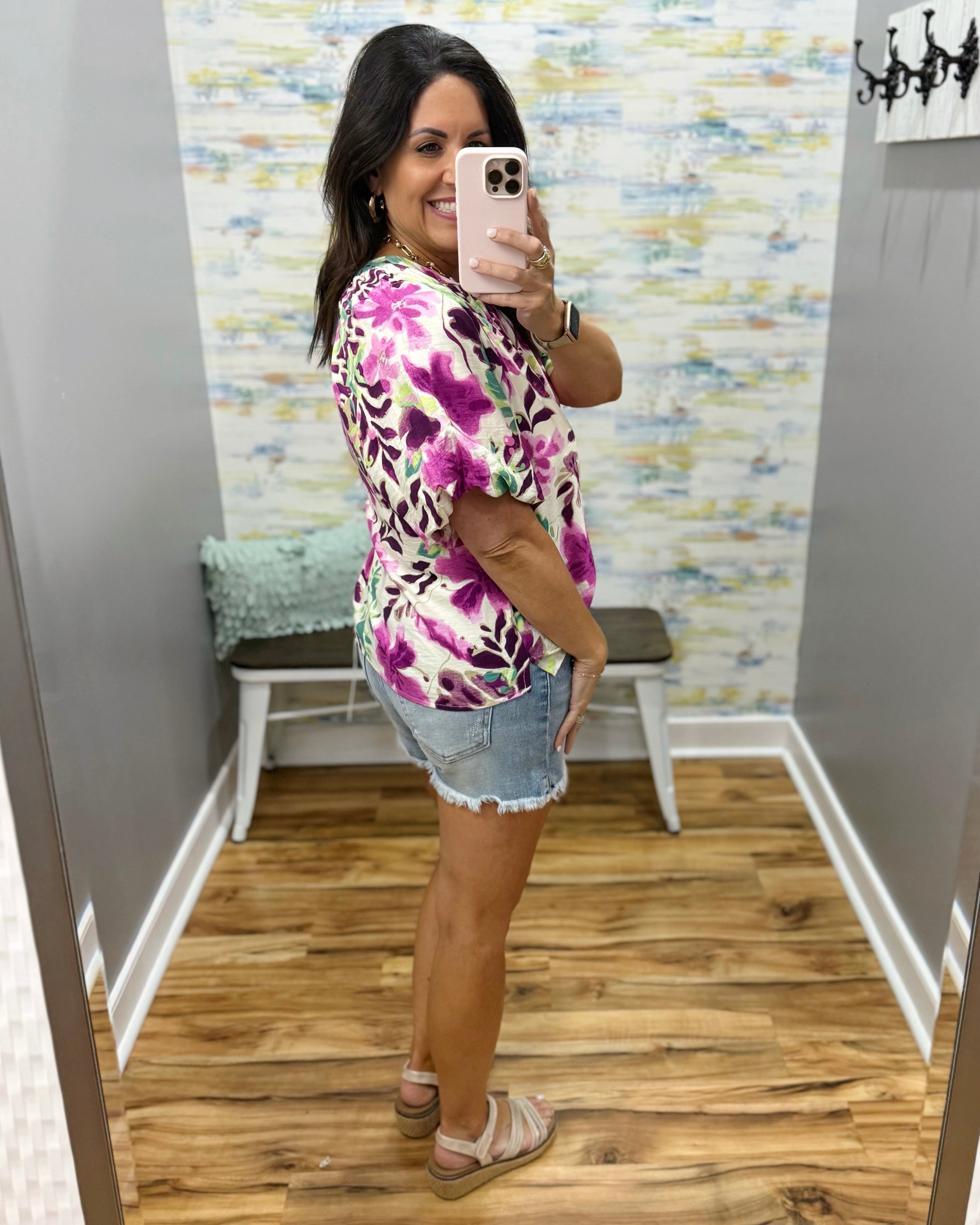 Woman taking a mirror selfie wearing a colorful floral shirt and denim shorts in a room with patterned wallpaper.