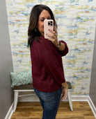 A woman takes a mirror selfie wearing a burgundy knit sweater and blue jeans in a room with a patterned wallpaper background.