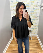 A woman takes a mirror selfie wearing a black short-sleeve pleated blouse and blue jeans in a fitting room with wood floors and a patterned wall.