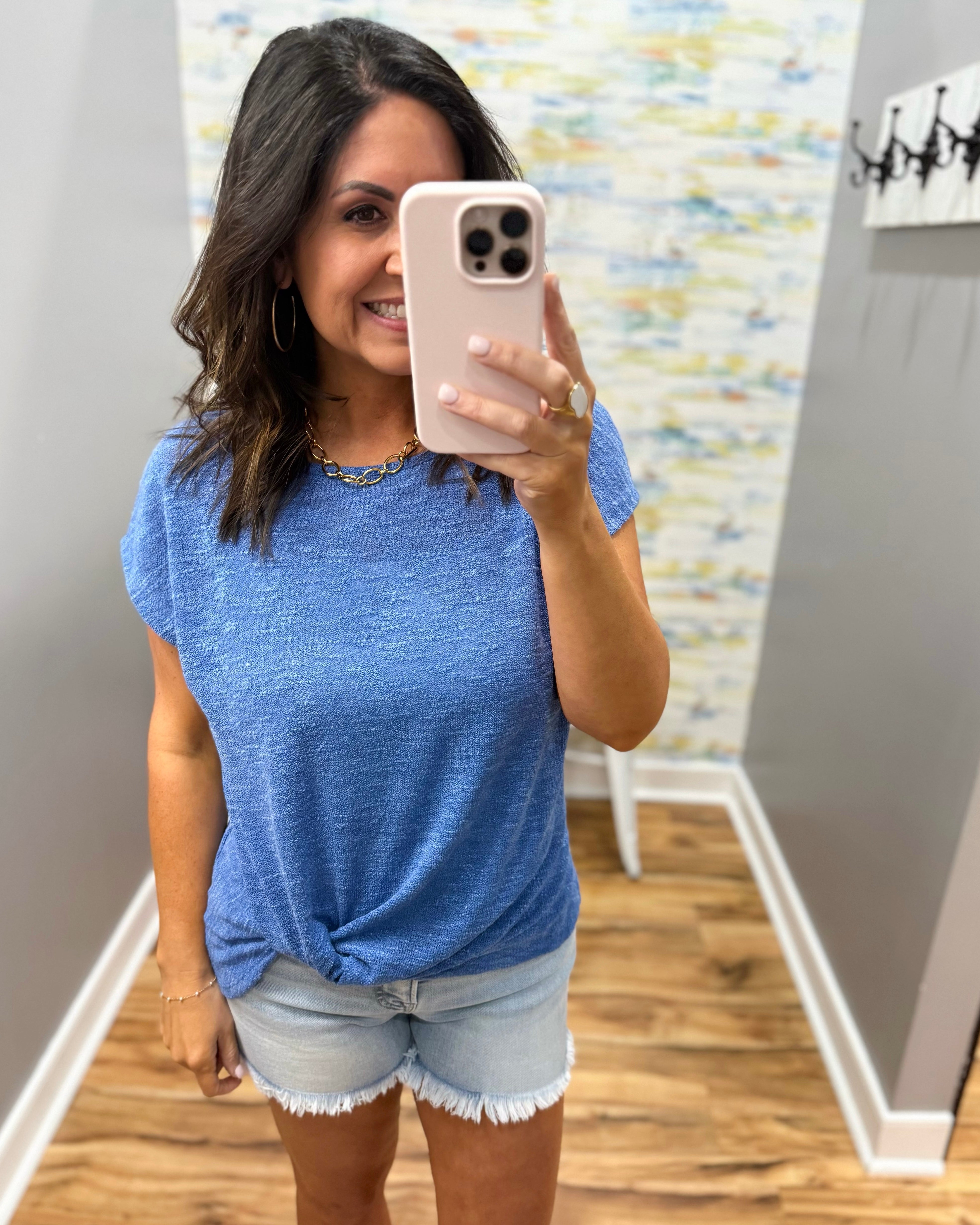Woman taking a mirror selfie wearing a blue top and denim shorts in a room with a patterned wall.