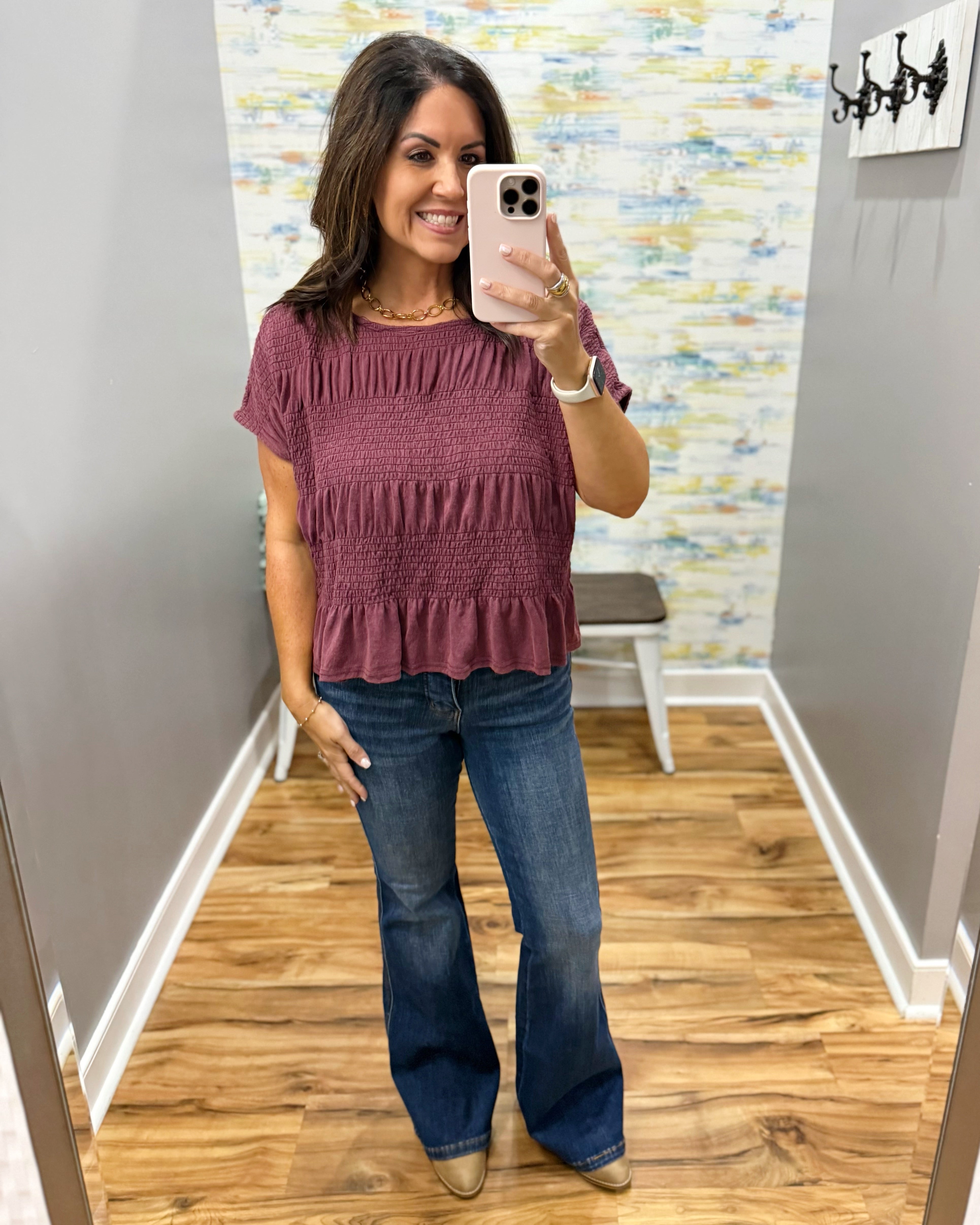 A woman takes a mirror selfie in a fitting room wearing a short-sleeve smocked ruffle top in dusty mauve and blue flare jeans.