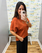 A woman takes a mirror selfie wearing a short-sleeve rust-orange knit top and black pants in a fitting room with a patterned wall and wood floor.