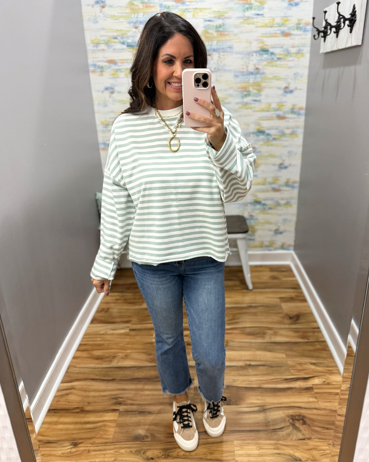 A woman takes a mirror selfie wearing a light green and white striped long-sleeve top, blue jeans, and beige sneakers with black laces in a fitting room.