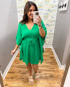 Woman wearing a green dress taking a mirror selfie in a room with wooden flooring and a colorful wall.