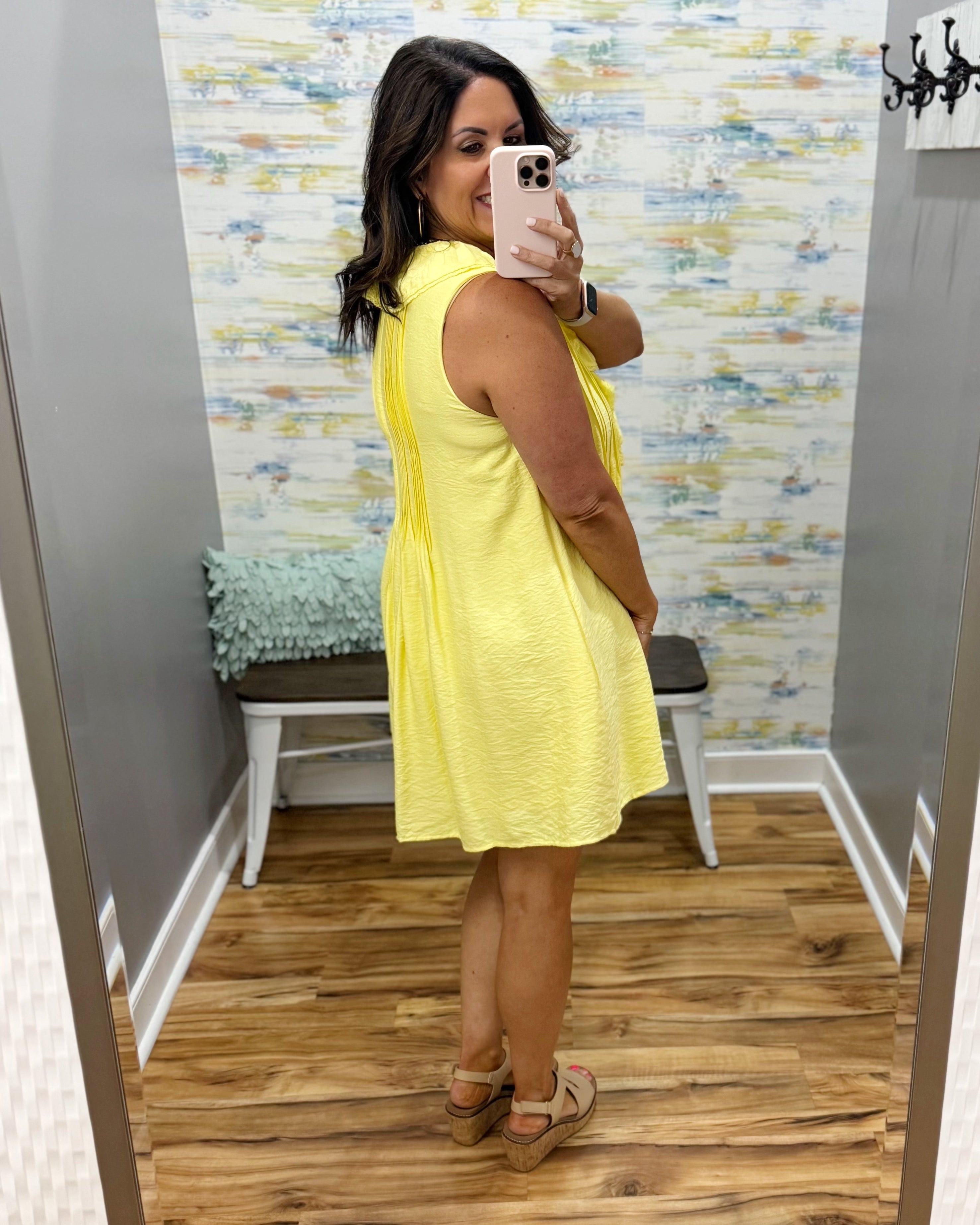 Woman taking a mirror selfie wearing a yellow dress in a room with patterned wallpaper.