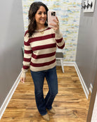 A woman takes a mirror selfie wearing a long-sleeve knit sweater with horizontal burgundy and cream stripes, paired with dark blue jeans and tan boots.
