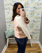 A woman takes a mirror selfie in a fitting room wearing a color-blocked knit sweater with cream, light pink, and taupe panels, paired with dark blue jeans.