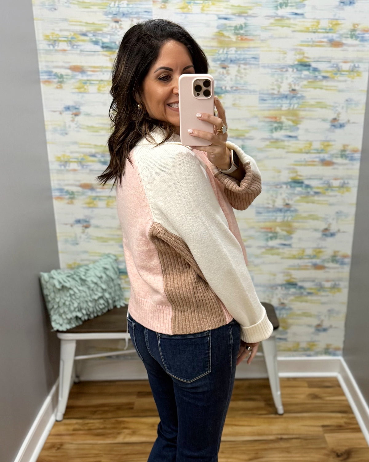 A woman takes a mirror selfie in a fitting room wearing a color-blocked knit sweater with cream, light pink, and taupe panels, paired with dark blue jeans.
