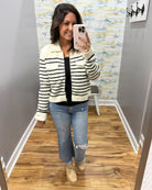 A woman takes a mirror selfie in a fitting room wearing a cream and black horizontally striped cropped zip-up jacket over a black top, blue distressed jeans, and beige lace-up boots.