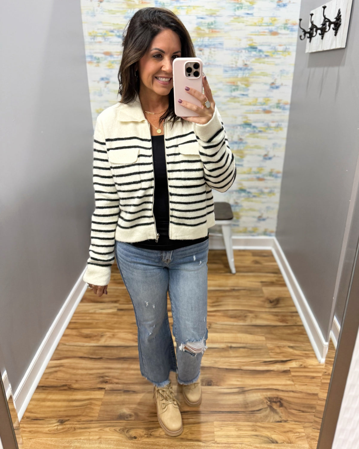 A woman takes a mirror selfie in a fitting room wearing a cream and black horizontally striped cropped zip-up jacket over a black top, blue distressed jeans, and beige lace-up boots.