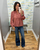 A woman takes a mirror selfie wearing a dusty rose long-sleeve blouse with a smocked chest and blue flare jeans in a fitting room.