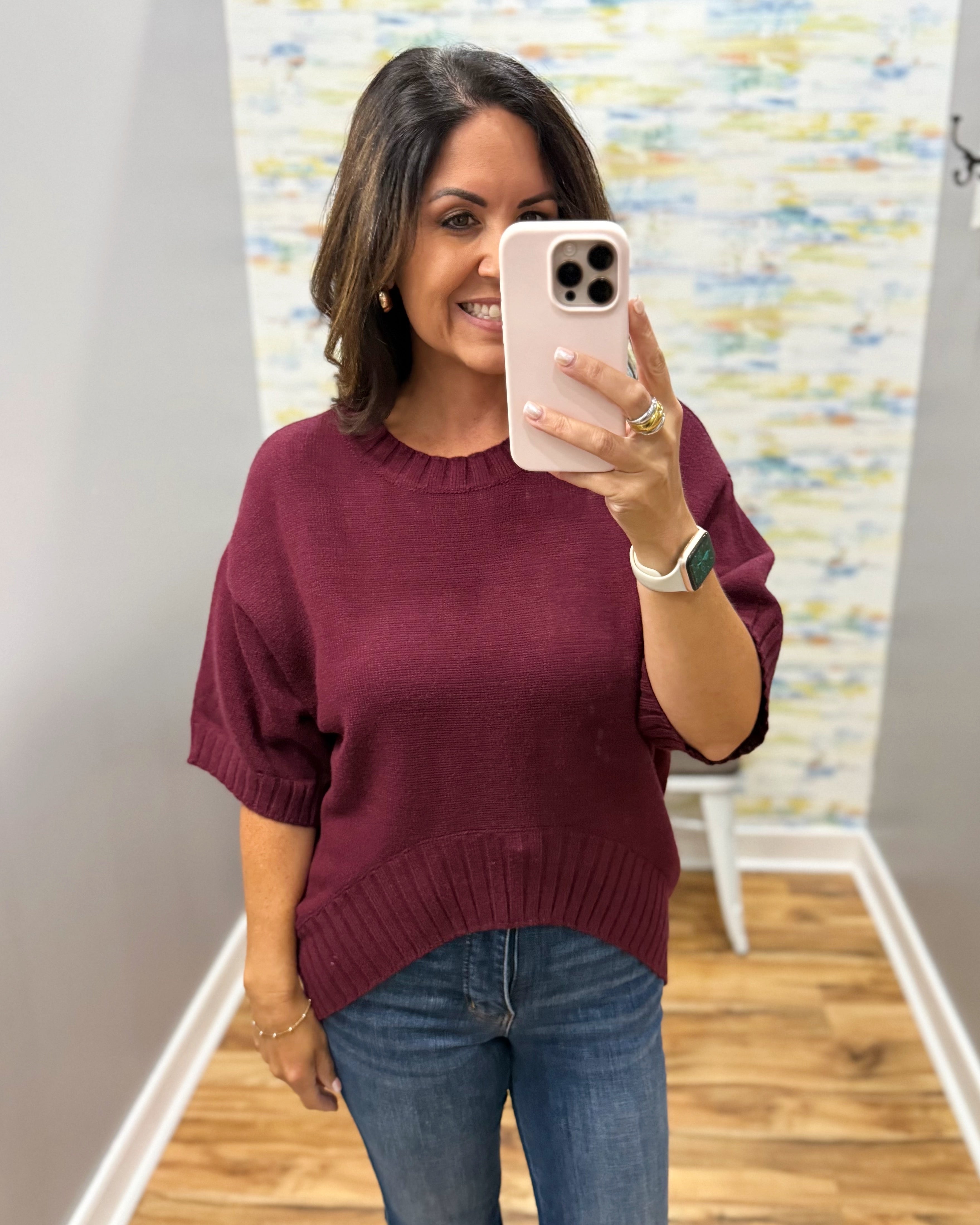 A woman taking a mirror selfie wearing a short-sleeve knit sweater in burgundy and blue jeans, holding a light pink smartphone.