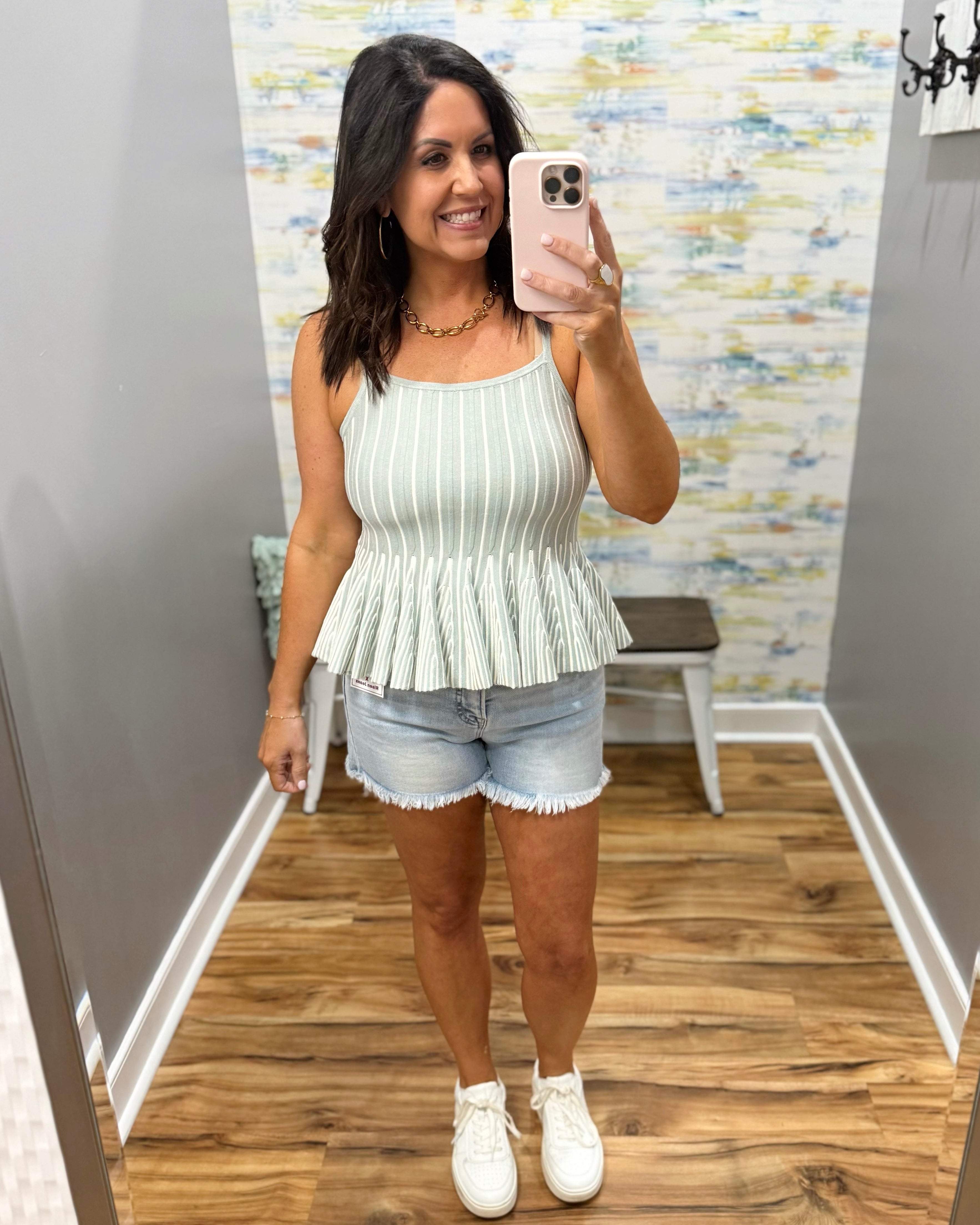 Woman taking a mirror selfie wearing a white pleated top and denim shorts in a room with a patterned wall.
