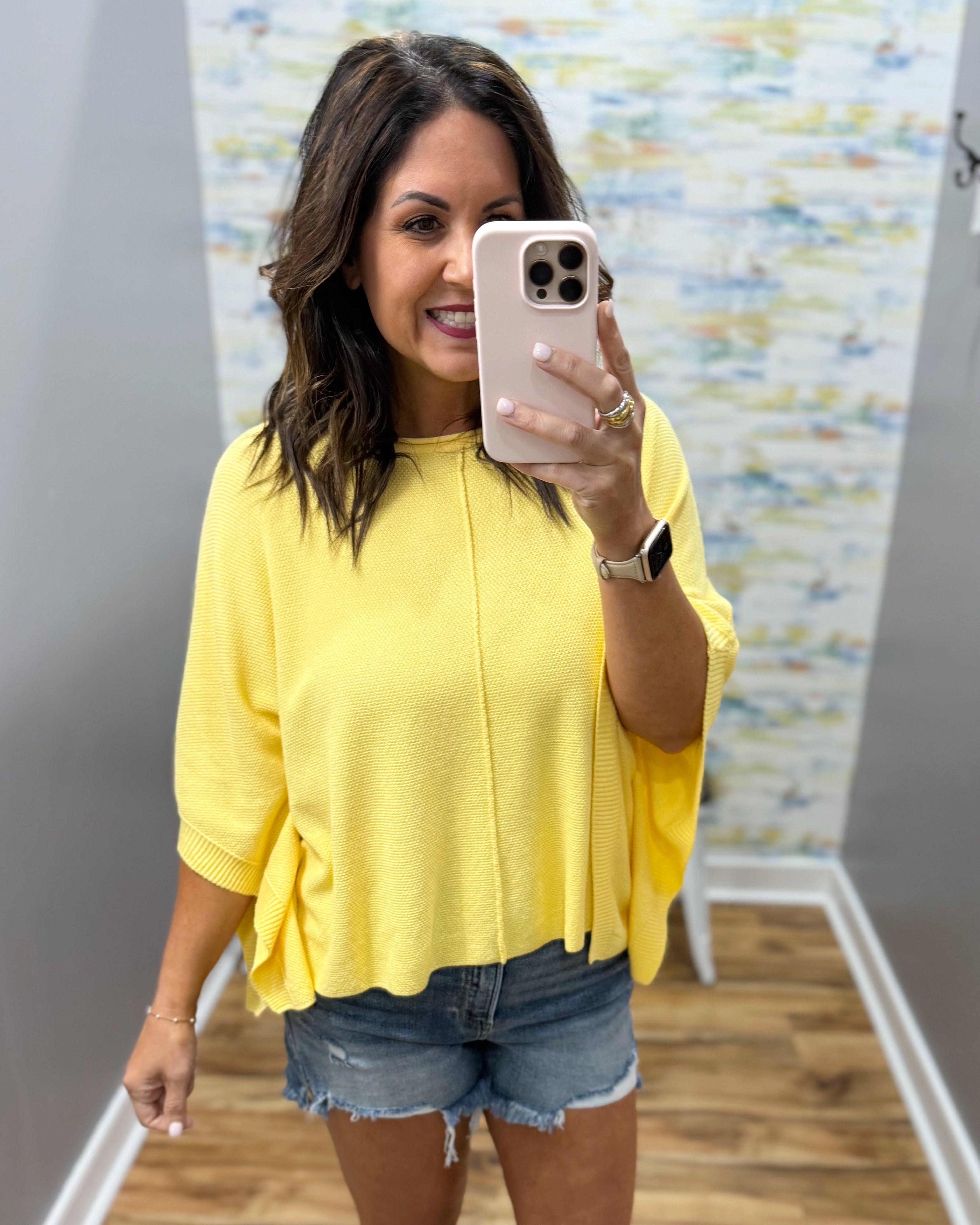 Woman taking a mirror selfie wearing a yellow top and denim shorts in a room with a colorful wall.