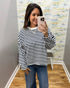A woman taking a mirror selfie wearing a navy and white horizontally striped long-sleeve top with blue jeans, standing in an indoor room with a wood floor and abstract wallpaper.