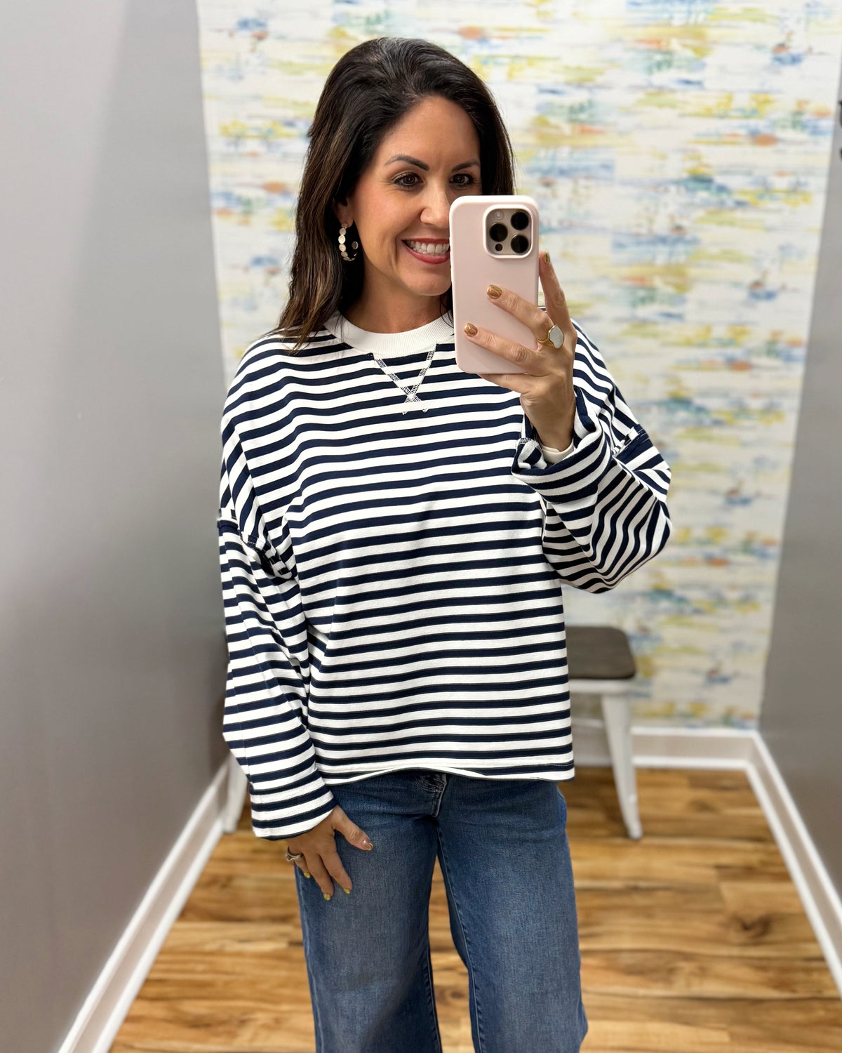 A woman taking a mirror selfie wearing a navy and white horizontally striped long-sleeve top with blue jeans, standing in an indoor room with a wood floor and abstract wallpaper.