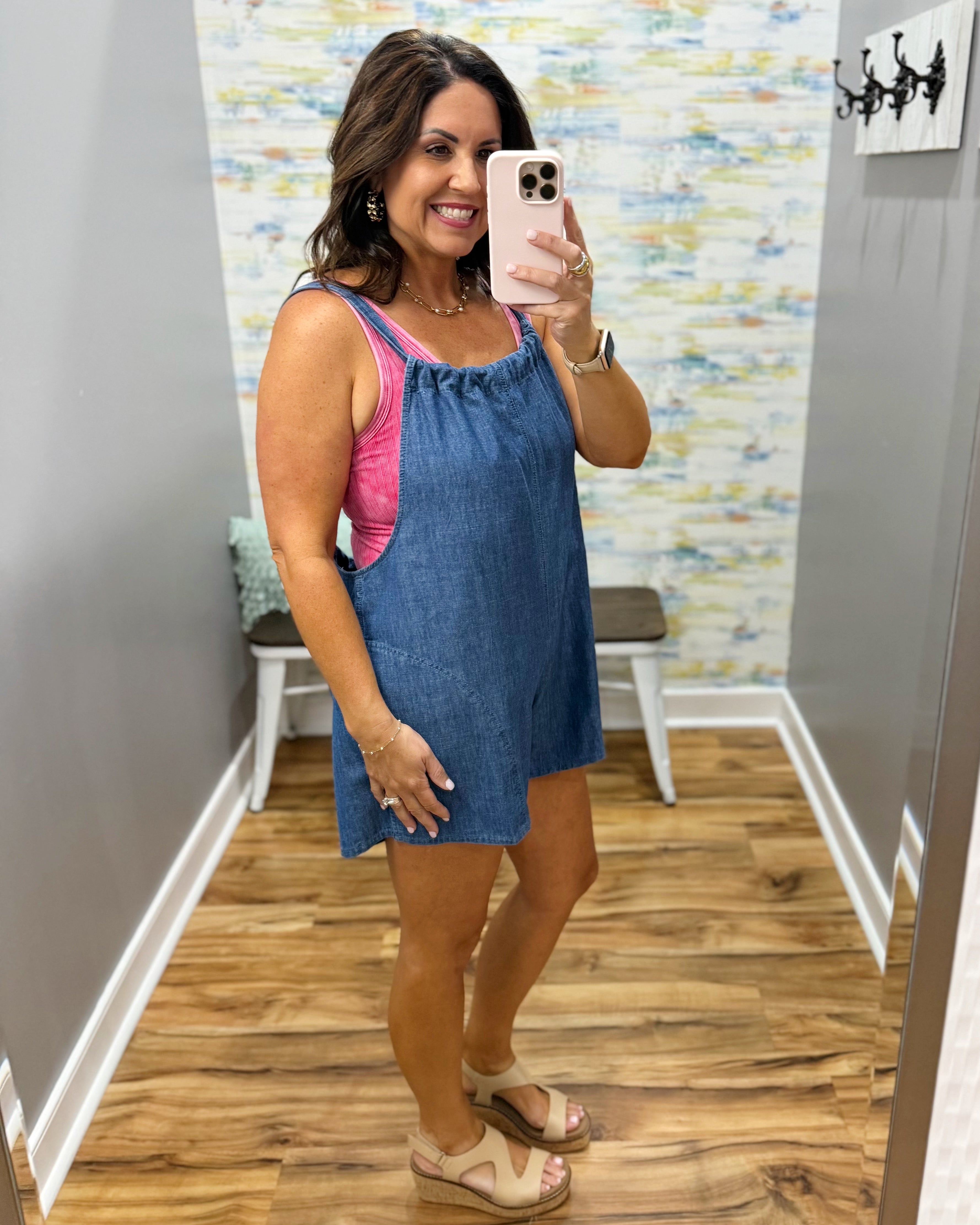 Woman taking a mirror selfie wearing a blue dress over a pink top in a room with a patterned wall.
