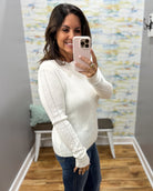 A woman takes a mirror selfie wearing a cream-colored knit sweater with textured cable detailing on the sleeves and dark blue jeans.