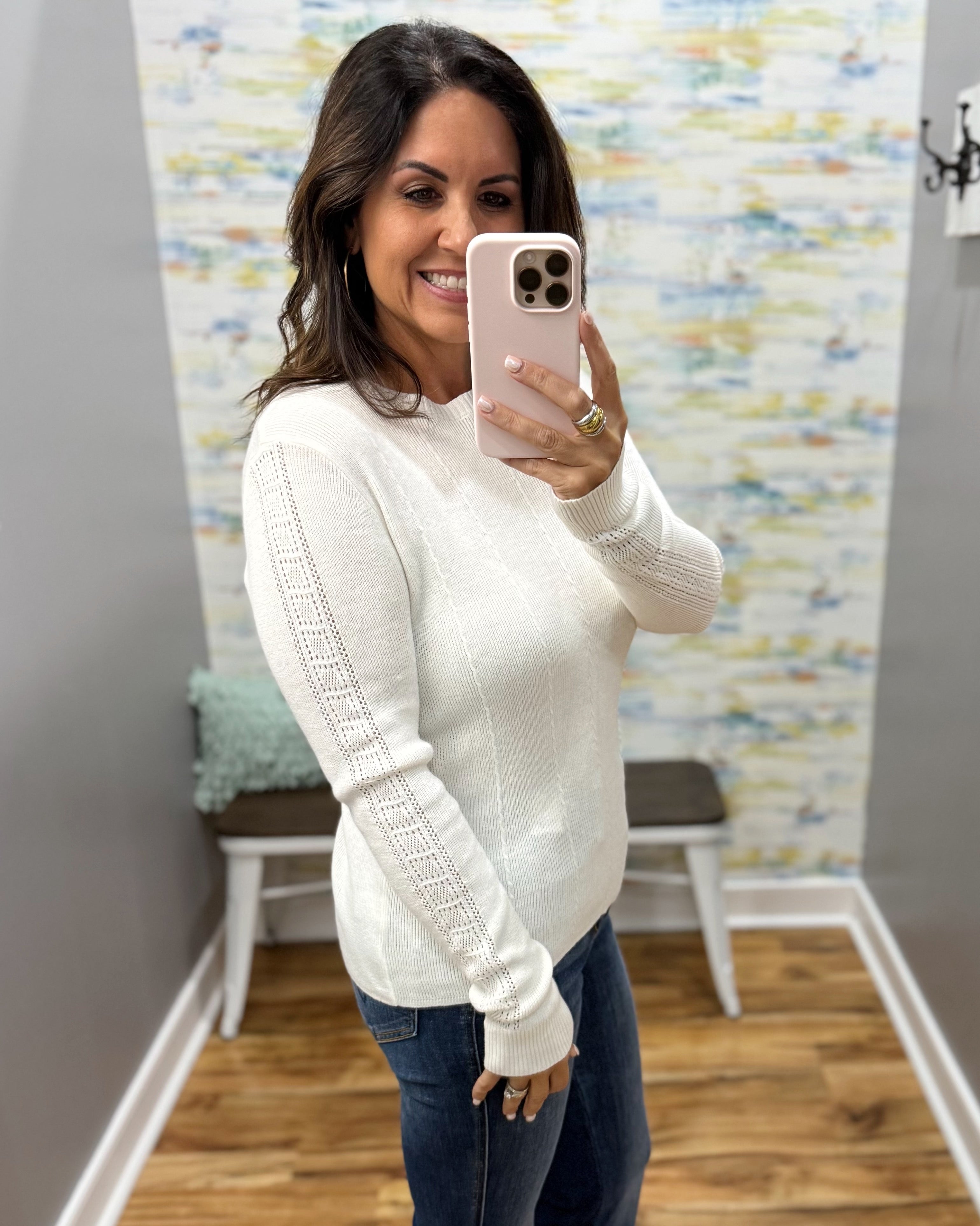 A woman takes a mirror selfie wearing a cream-colored knit sweater with textured cable detailing on the sleeves and dark blue jeans.