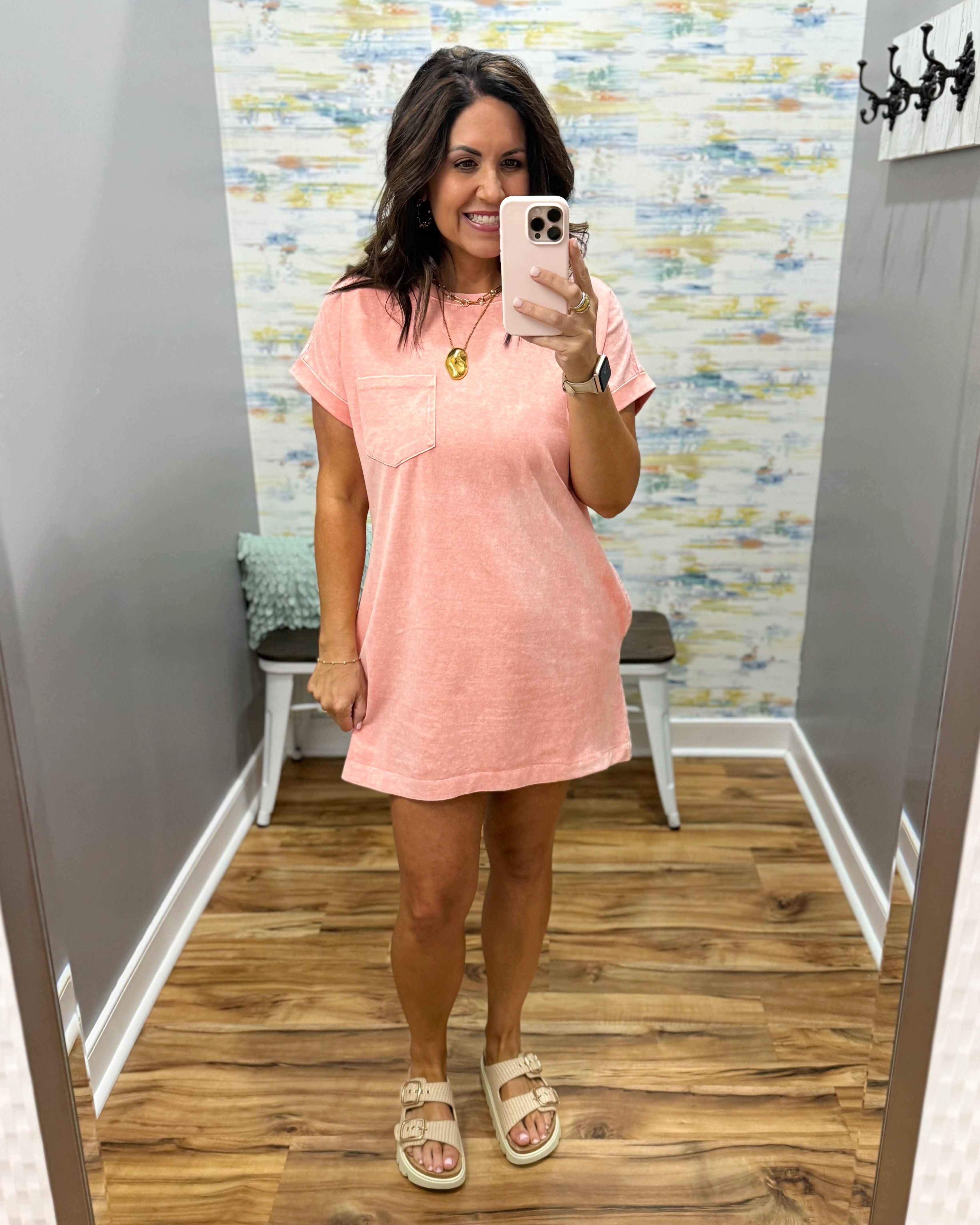 Woman taking a mirror selfie wearing a pink dress in a room with a patterned wall.