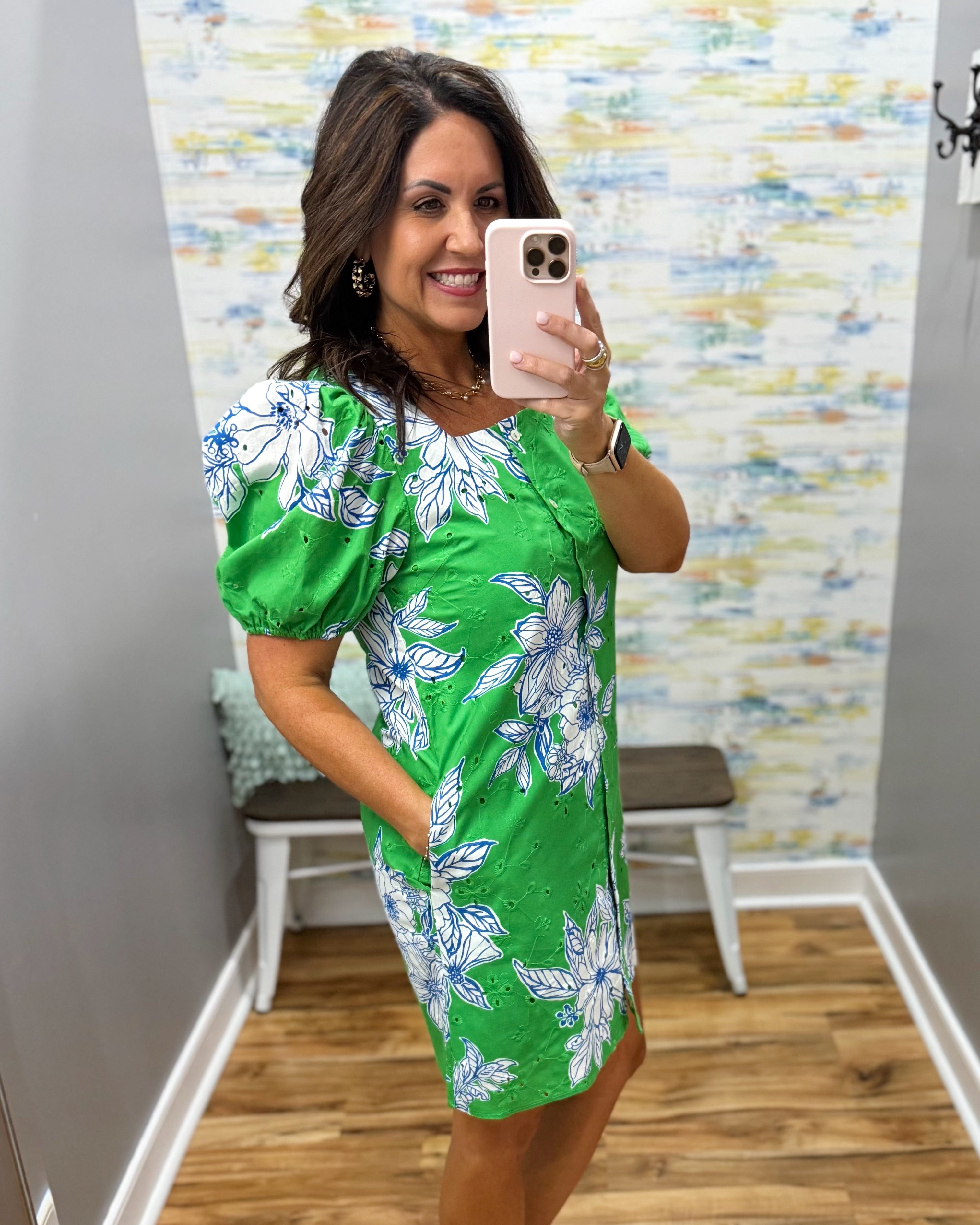Woman taking a mirror selfie wearing a green floral dress in a room with a colorful wall.