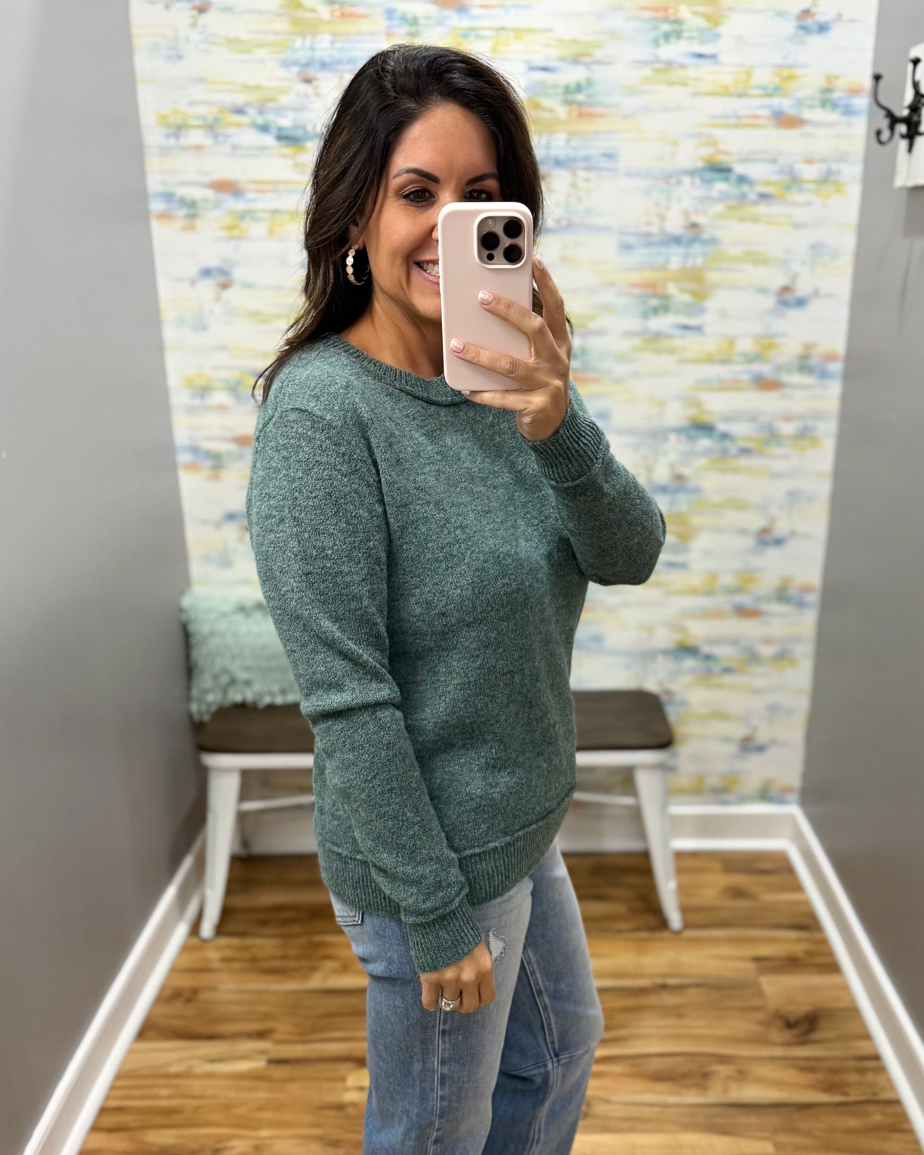 A woman takes a mirror selfie wearing a heathered sage green long-sleeve knit sweater and light blue jeans in a fitting room with a wood floor and abstract wallpaper.