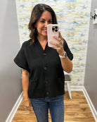 A woman takes a mirror selfie wearing a black short-sleeve button-front top and blue jeans in an indoor room with gray walls and wood flooring.