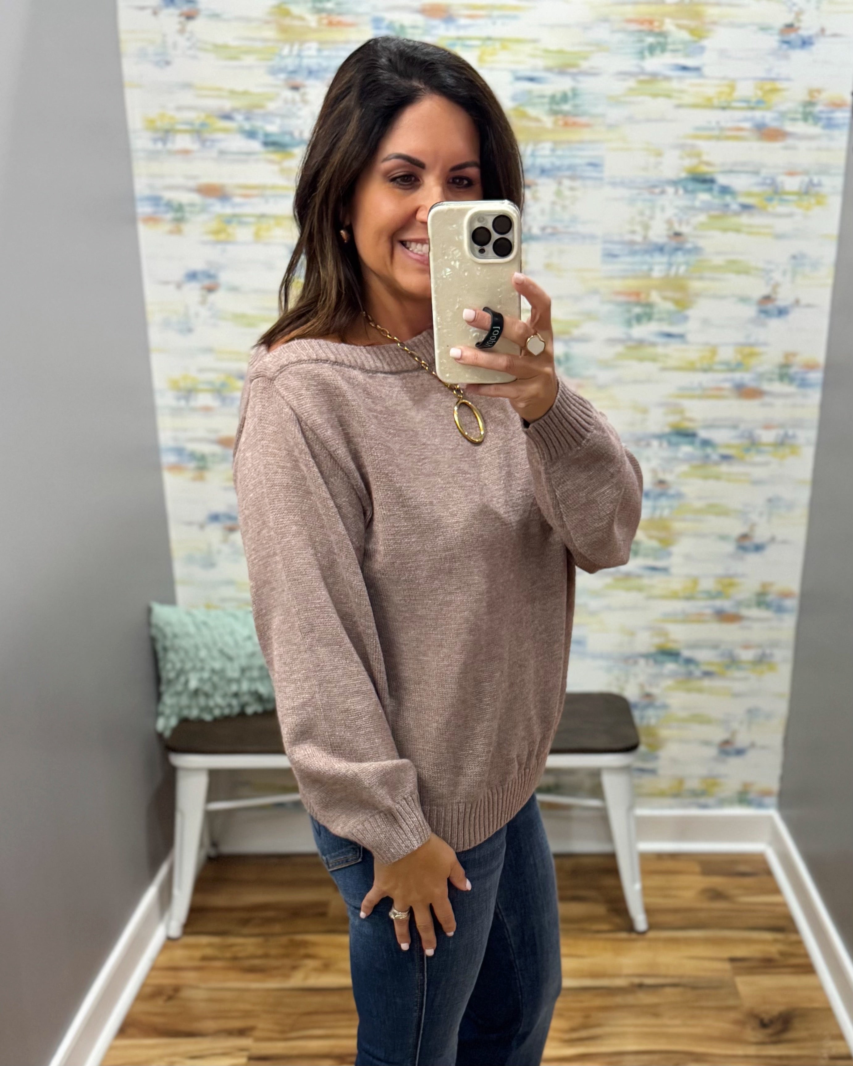 A woman takes a mirror selfie wearing a loose, dusty rose knit sweater and dark blue jeans in a room with a patterned wallpaper background.