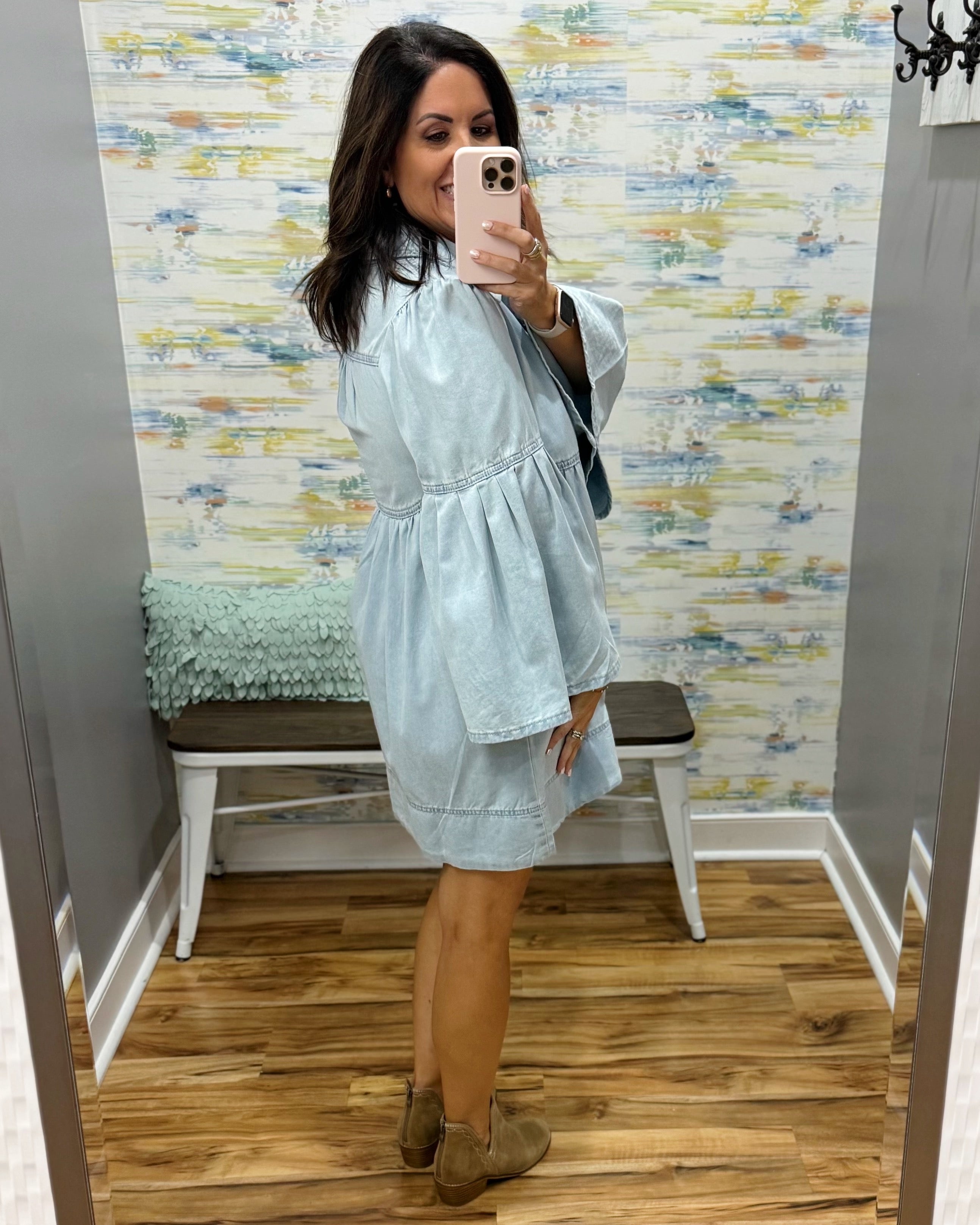 A woman takes a mirror selfie in a fitting room wearing a light blue short dress with a gathered waist and flared sleeves, paired with brown ankle boots. The background features a patterned wallpaper, a wooden bench, and a wood floor.