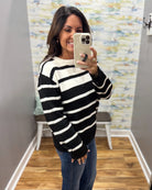 A woman takes a mirror selfie wearing a black and white horizontally striped knit sweater and blue jeans in a fitting room with wood floors and a patterned wallpaper background.