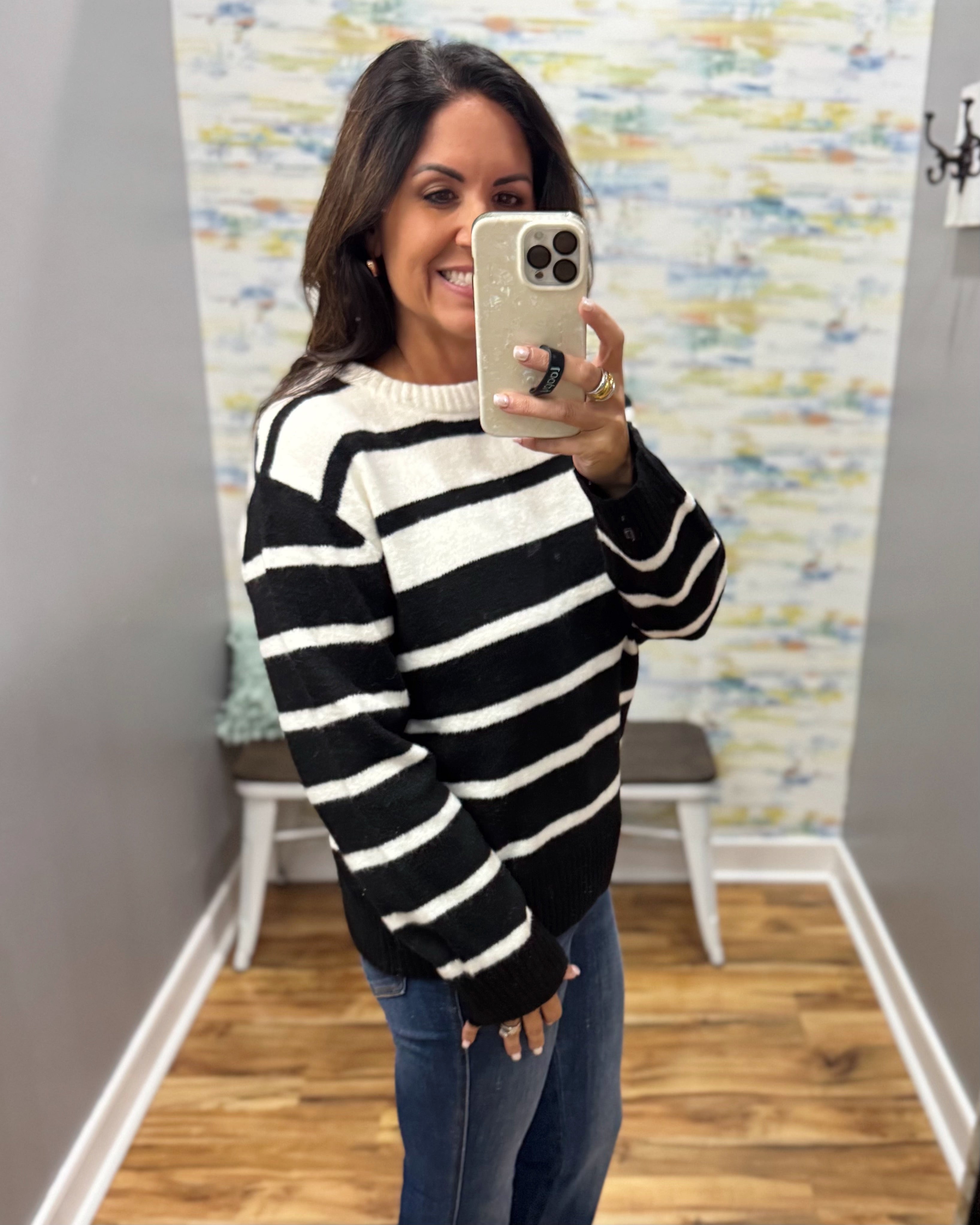 A woman takes a mirror selfie wearing a black and white horizontally striped knit sweater and blue jeans in a fitting room with wood floors and a patterned wallpaper background.