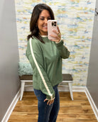 A woman takes a mirror selfie wearing an olive green long-sleeve sweatshirt with cream-colored stripes down the sleeves, paired with blue jeans.